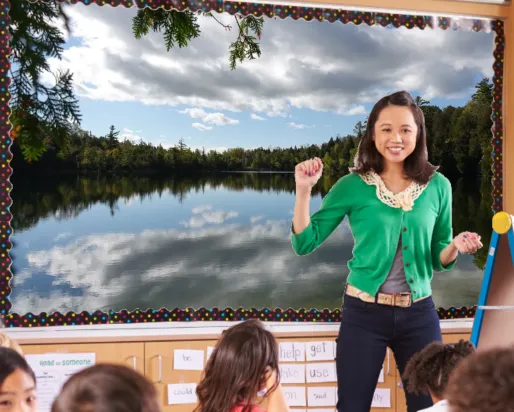 A teacher in a green cardigan stands in front of a smartboard at the front of a class. The board displays an image of a glassy lake.