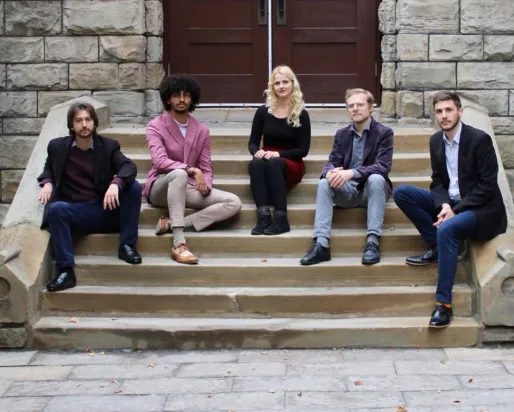 Five young people sit facing the camera on the stone steps of a historic building.