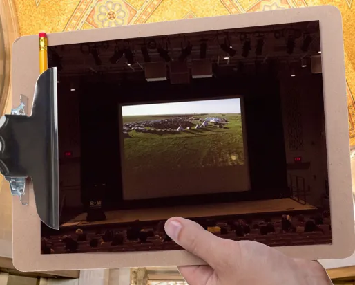 A hand holds a clipboard in front of a large gallery with a gold mosaic ceiling. The clipboard displays a picture of students in a darkened theatre watching a presentation.