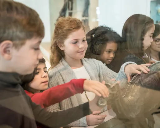 Several young students in a diverse class touching a bronze statue of a maiasaur.