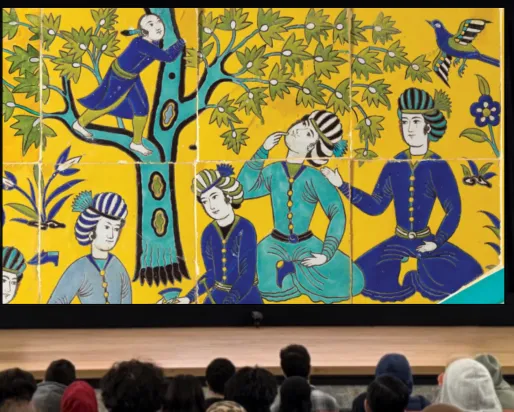 A woman stands behind a podium on a stage. The screen behind her displays an image of a colourful Islamic tile depicting several people at a picnic.