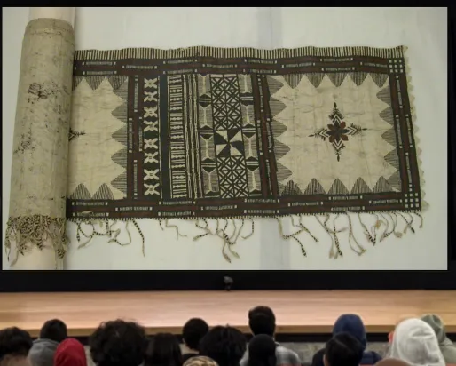 A smiling Indigenous woman stands behind a podium on a stage. On a screen next to her is an image of a length of cloth with intricate patterns in dark black and brown ink.
