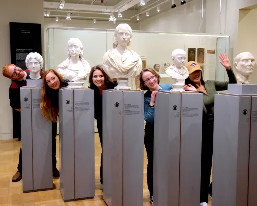 “A group of people playfully pose by leaning out between classical marble busts in a museum gallery.