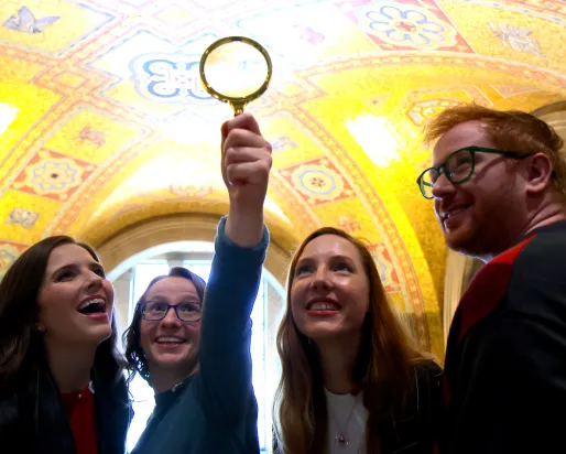A group of people smile and hold up a magnifying glass while exploring the ornate golden ceiling of the museum rotunda.