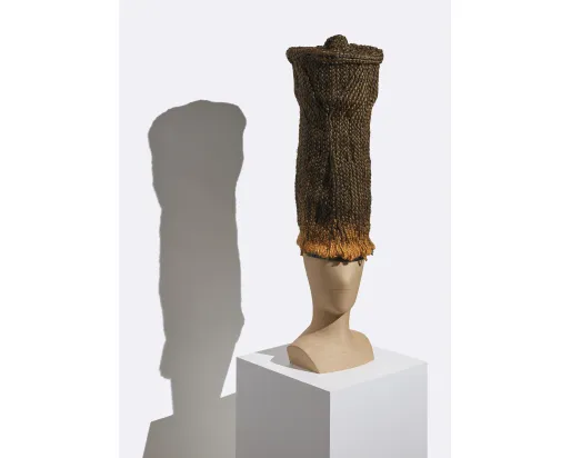 Tall cylindrical woven basket with a flared rim displayed on a white pedestal casting a shadow on the wall behind.