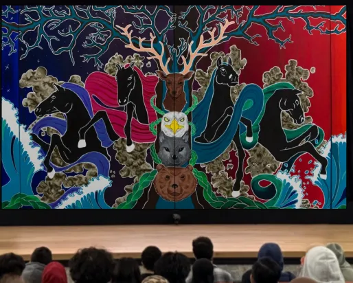 An Indigenous woman stands on a stage behind a podium. A screen behind her displays a painted mural.