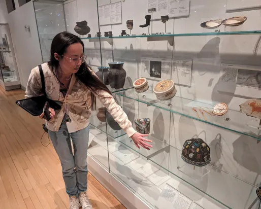 An Indigenous woman with long, dark hair and glasses stands by a museum case filled with belongings showing beadwork, quillwork, and other artistic traditions.