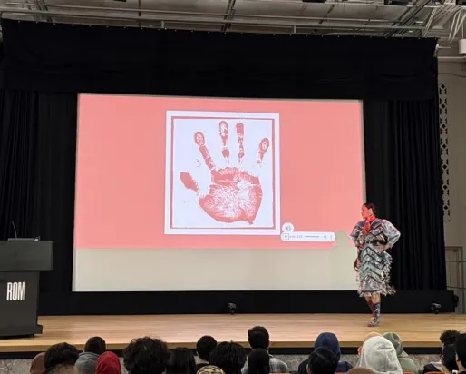 An Indigenous woman wearing a jingle dress stands on a stage in a theatre. The screen behind her shows a white handprint on a red screen.