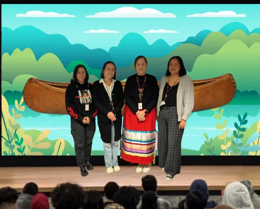 Four Indigenous women stand on a stage. The screen behind them displays an image of a canoe on an illustrated river.