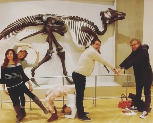 A group of people poses playfully in front of a dinosaur skeleton, arranging their bodies to spell out shapes.