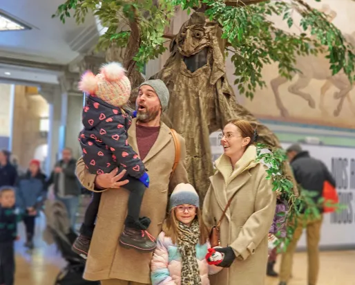 Two adults and two children stand indoors near a large tree-like display; one adult holds a bundled-up toddler while the others smile, with people walking in the background.