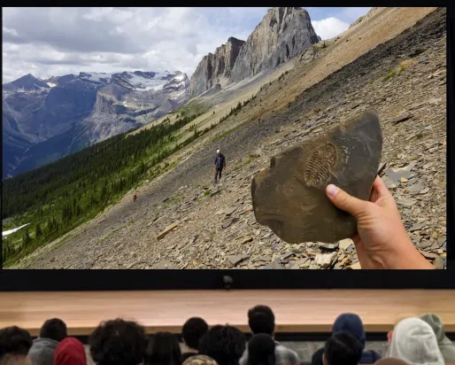 A man wearing a hard hat with goggles stands behind a podium on a stage. A screen next to him depicts a shale-covered hillside with mountains in the background. A lone figure walks across the hillside. A hand holds up a fossil in the foreground.