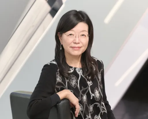 Asian woman with glasses wearing a black dress with a white floral pattern, sitting on a black chair against a modern background.