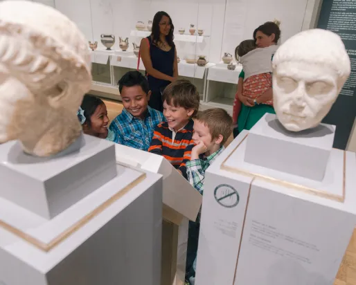 Two adults watch a smiling group of children in front of a group of marble busts