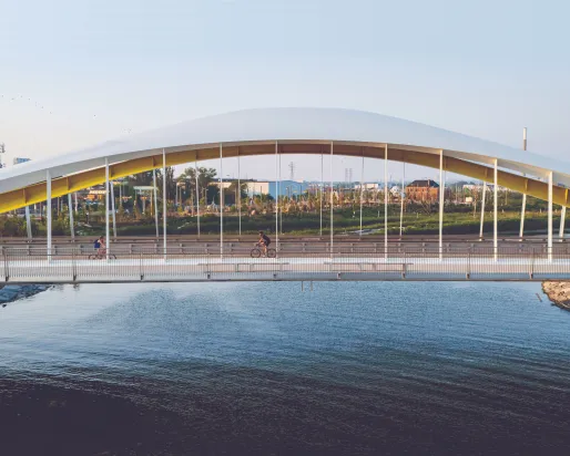 Wide view of a modern arched pedestrian bridge spanning a body of water, with cyclists crossing beneath a curved white canopy supported by vertical cables, set against a city landscape and pale blue sky in warm evening light.
