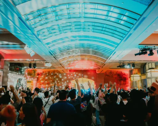 Crowd of people in a large indoor venue with a high curved glass ceiling, raising their arms toward a brightly lit stage. Colorful red and blue lighting and spotlights illuminate the space, with artwork and event lighting visible along the walls