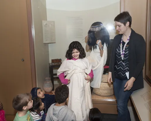 A museum teacher dresses a smiling child in an Egyptian dress as classmates watch.