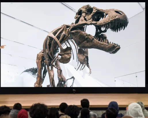 A man stands at a podium on a stage in front of an audience of students. He points at a dinosaur on a screen.