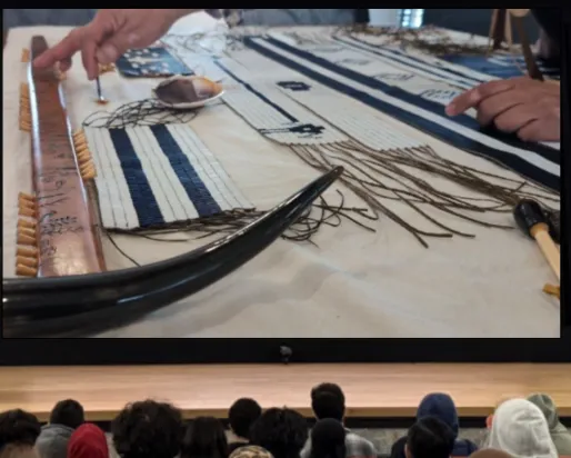 Une femme autochtone se tient debout derrière un pupitre, sur une scène, face à un groupe de jeunes, et désigne d'un geste les ceintures de wampum et d'autres objets exposés sur un écran.