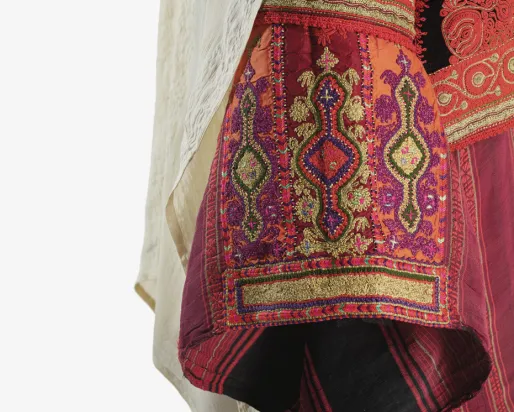 Gros plan sur un vêtement traditionnel orné de broderies complexes aux tons rouges, orange, dorés et violets, représentant des motifs géométriques et floraux. Un tissu drapé de couleur claire tombe le long de ce textile à la texture riche, mettant en valeur le savoir-faire artisanal et les coutures décoratives.