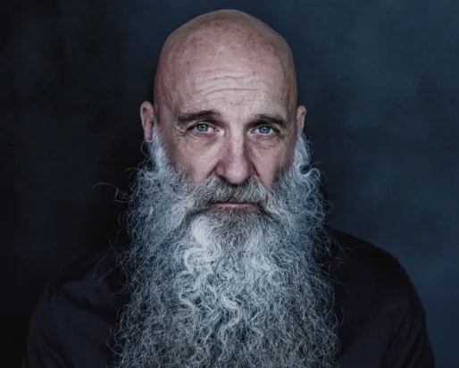 Portrait en studio d'un homme chauve, portant une longue barbe blanche et une chemise sombre boutonnée, sur fond bleu foncé.