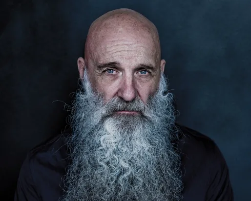 Studio portrait of a bald individual with a long white beard, wearing a dark button-up shirt against a dark blue background.