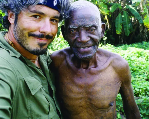 Un homme vêtu d'une chemise verte et d'un bandana bleu prend un selfie avec un maître tatoueur Makondé.
