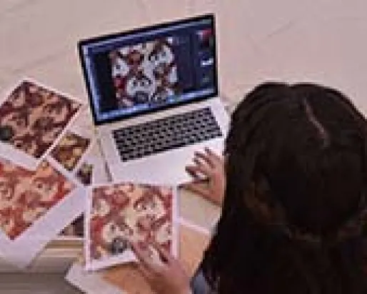 A young woman sits in front of a laptop computer surrounded by squares of patterned fabric.
