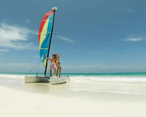 A man & a woman in bathing suits pose on a small catamaran on a sun-lit, white sand beach
