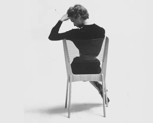 Monochrome 1950s image of the back of a woman in a black dress seated on a chair made of a wood frame and cord strung seat and back.