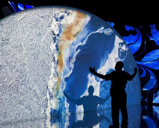 Projection of a planet with a silhouette of a man with his hands in the air looking at a large projection of a planet