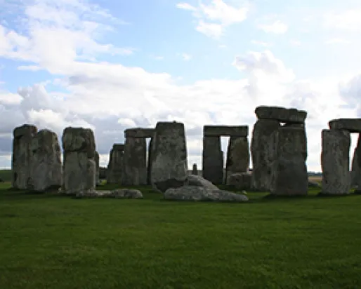 Large rectangular stone pillars in a ring in a field of green grass on a partly cloudy day. Large rectangular rocks placed horizontally on top span between some pairs of pillars. Farm fields are visible in the background.
