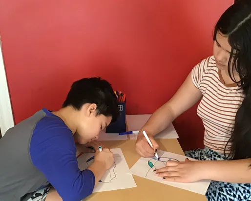 wo girls, one with short dark hair and the other with long dark hair, sit at a table and colour iceberg drawings with markers.
