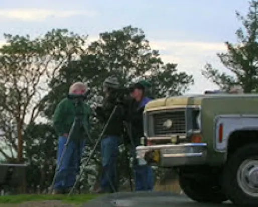 Three people standing together next to the front of a motor vehicle, with leafy green trees in the background.
