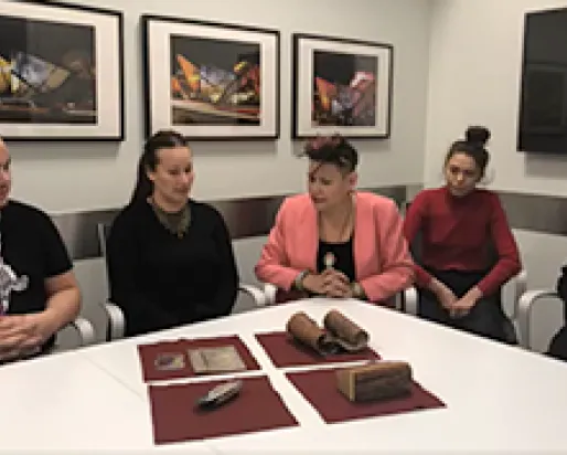 Five people sit around a white table in a meeting room with brown objects on the table.
