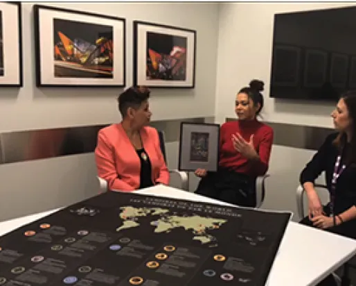Three people sit around a table. On the table is a map showing stories of blood-feeding creatures and their origins around the world. Thée Wind Dancer holds a framed picture of the Wild Woman of the Wood.
