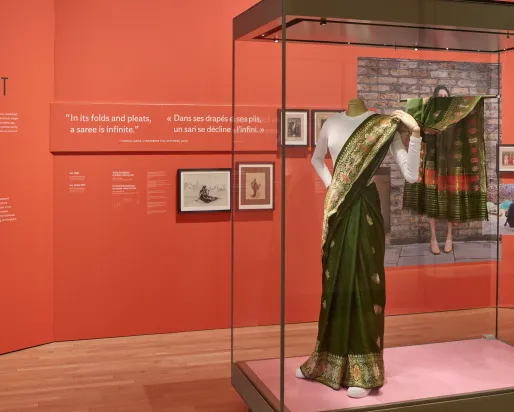 Sari, 1950s Installation showing red text panels behind a glass case containing a green silk sari with woven metallic thread worn over a white T-shirt, shown on a mannequin wearing white running shoes.