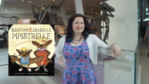A woman in a dinosaur dress stands in a gallery of dinosaur skeletons. Next to her is the cover of the book "Burton & Isabelle Pipistrelle."