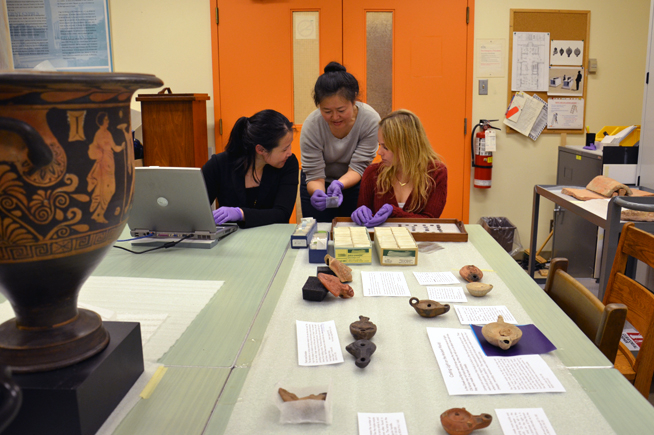 The ROM's Kay Sunahara examining Roman coins with interns Menghan Yan & Clare Schwartzberg, surrounded by artifacts being prepared for the upcoming Ancient Greece & Rome Weekend exhibit