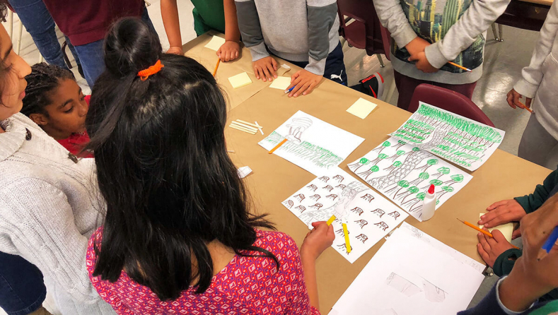A small group of elementary school students stand around a table, as one student shows sheets of paper with forest drawings on them.