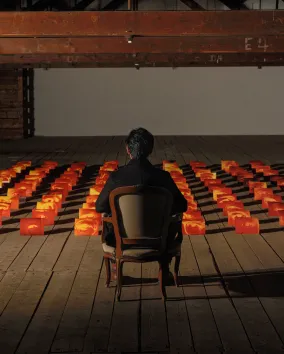 A person sits alone in a wooden chair facing an art installation of glowing orange and red blocks arranged in a grid, each block featuring a human eye, set in a dimly lit industrial-style room with exposed beams and wooden flooring.