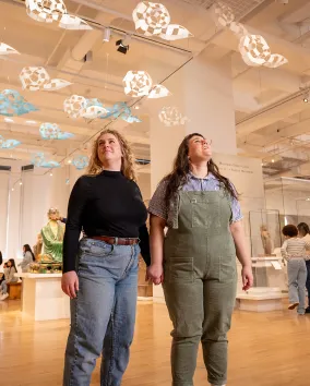 Two visitors hold hands while looking up at a hanging art installation in a museum gallery.