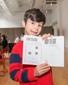 Boy in a red and blue striped sweater holding a white paper booklet with a QR code and text, indoors at an event.