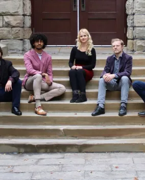 Five young people sit facing the camera on the stone steps of a historic building.