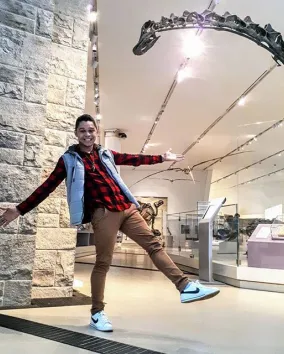 A person smiling and posing with arms outstretched in a museum gallery, standing beneath the long curved neck of a large dinosaur skeleton display.