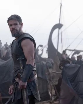 A bearded warrior in dark leather armor stands on a misty shoreline with an axe at his side, while ancient Greek-style ships and a busy encampment fill the background.