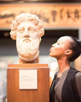 A girl leans toward a large marble bust in a museum as if giving it a kiss, with ancient artifacts and softly lit exhibits visible in the background.