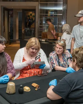Four people seated at a table examining fossilized skulls, with visitors and a museum entrance in the background.