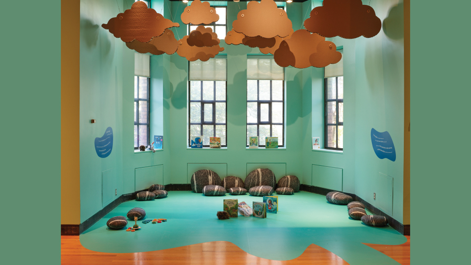 View of a windowed, reading alcove painted in calming green tones, with cloud cutouts suspended from above and lounging pillows that resemble rocks and boulders laid on the floor.