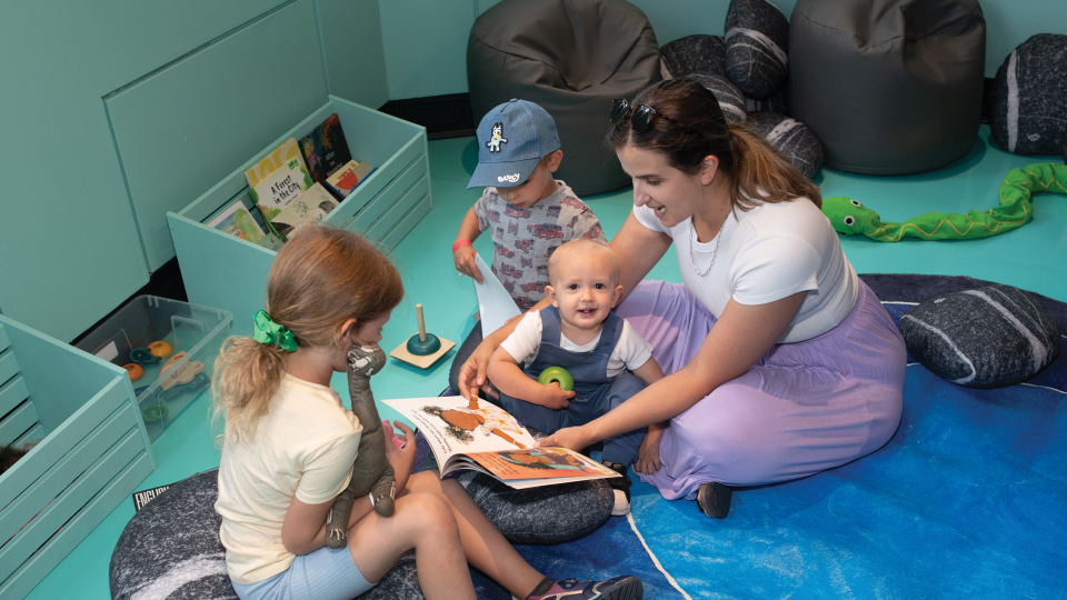 An adult sits on a floor mat, reading a picture book to a baby, while two other small children look on.